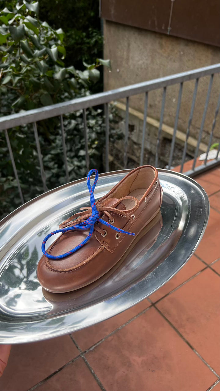Boat Loafer Brushed Brown von Posa auf silbernem Tablett im Freien, mit blauer Schnürung und Balkonkulisse.
