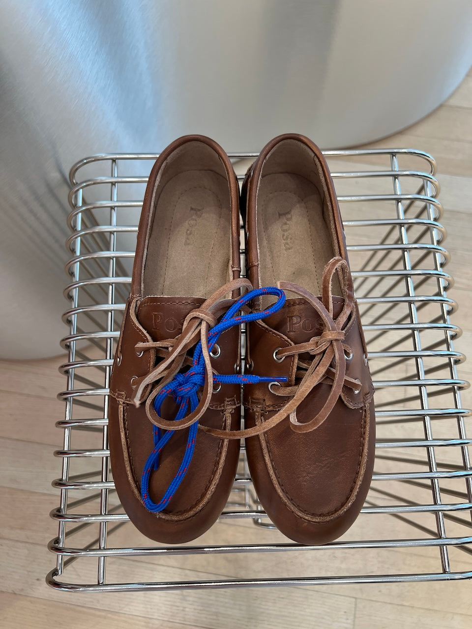 Posa Boat Loafer Brushed Brown von oben auf einem Metallgitter, mit blau-roter Ersatzschnürung drapiert.